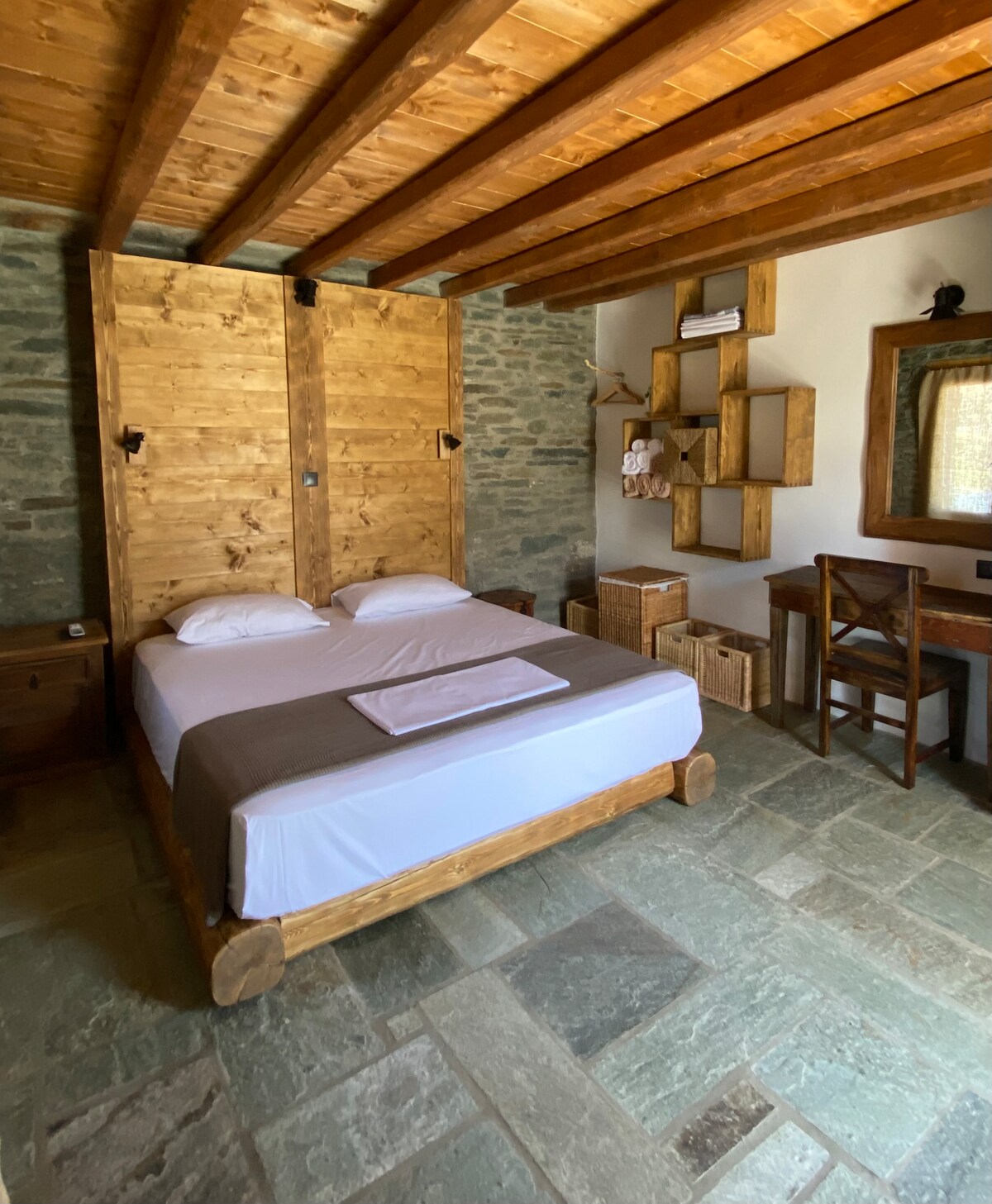 A bedroom features a double bed with a simple white duvet and a brown throw. Natural stone walls complement wooden accents, including sliding barn doors and shelving. A small desk and chair are positioned against one wall, enhancing the functional layout of the space.