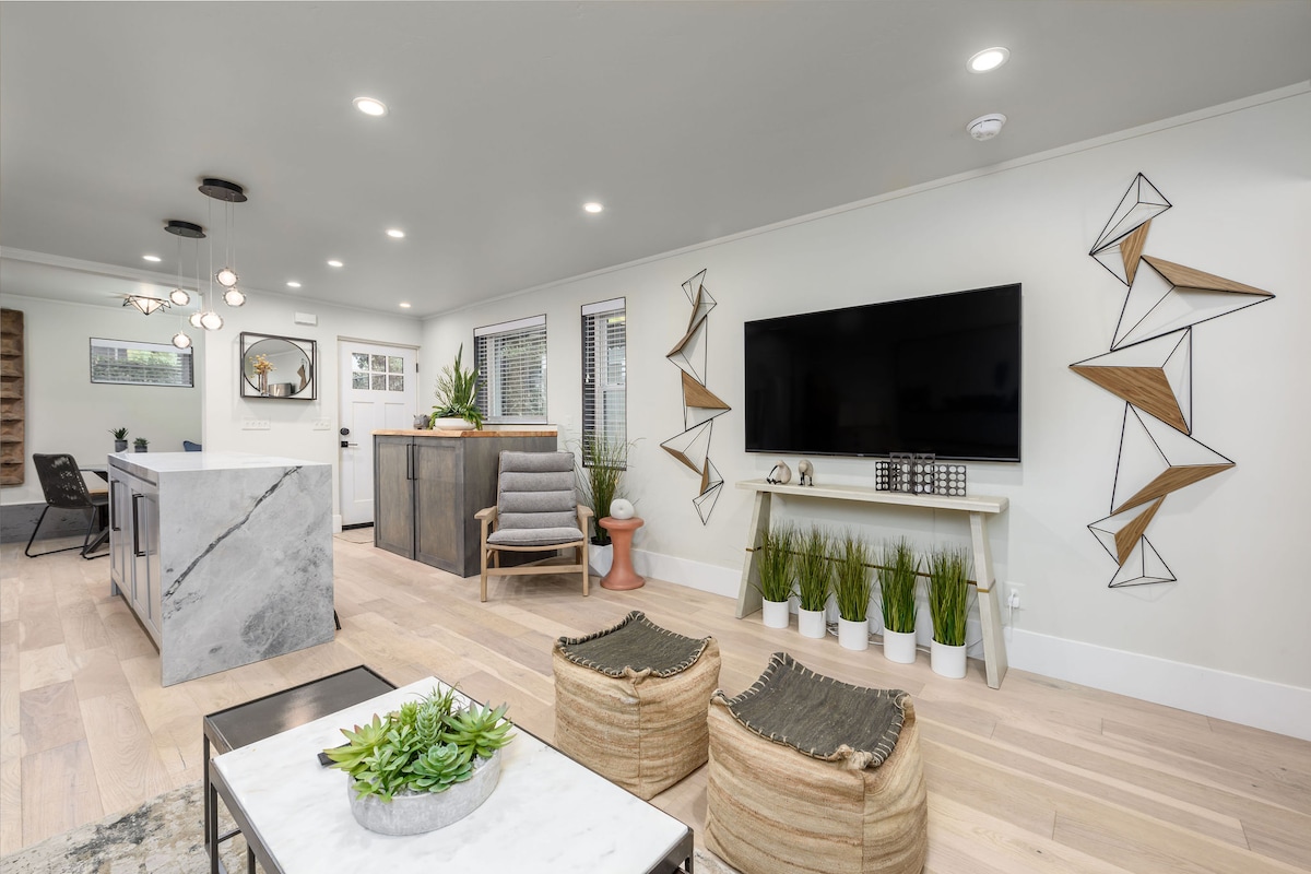 An open living space features a large television mounted on a light wall, surrounded by decorative geometric wall art. A gray kitchen island with seating is visible, along with potted plants and two stylish, woven poufs that enhance the contemporary decor.