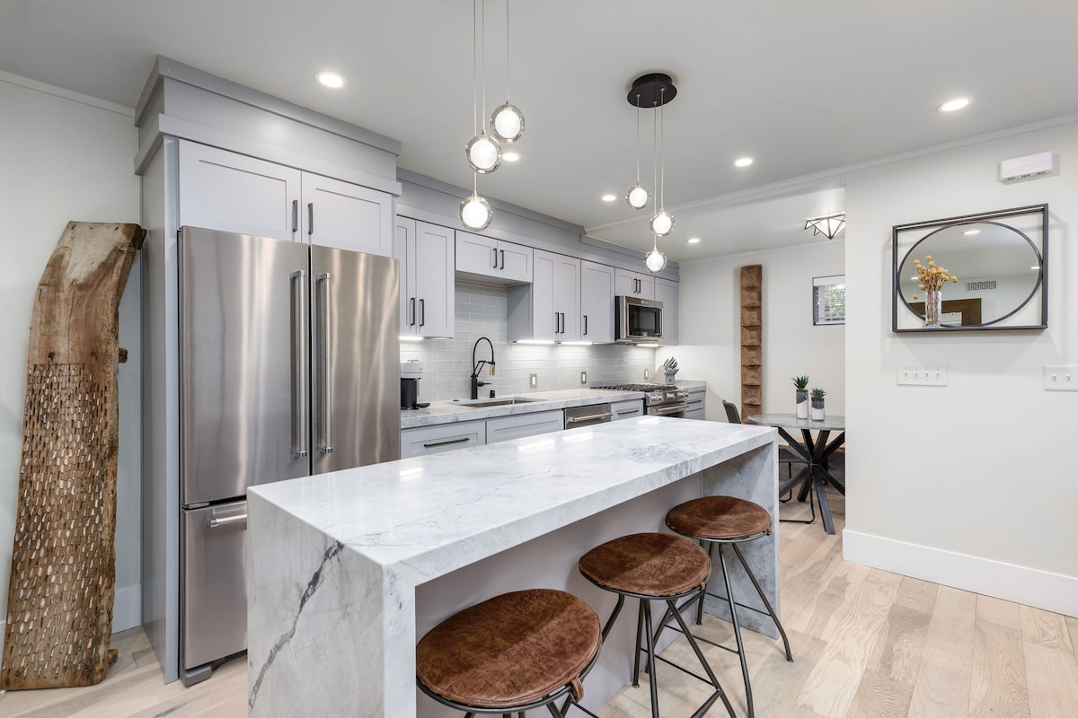 A modern kitchen is featured with sleek light gray cabinetry and stainless steel appliances. A large marble island provides ample space for meal preparation, complemented by three round, wooden bar stools. Soft lighting fixtures hang above, enhancing the functional layout.
