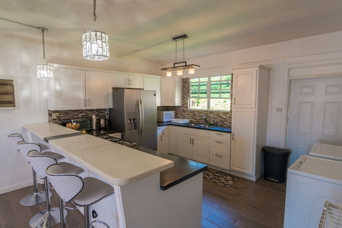 A modern kitchen features white cabinetry and stainless steel appliances, including a refrigerator and microwave. A countertop with four bar stools is positioned in front of a large window, allowing natural light to illuminate the space. Subtle tile accents add texture to the room.