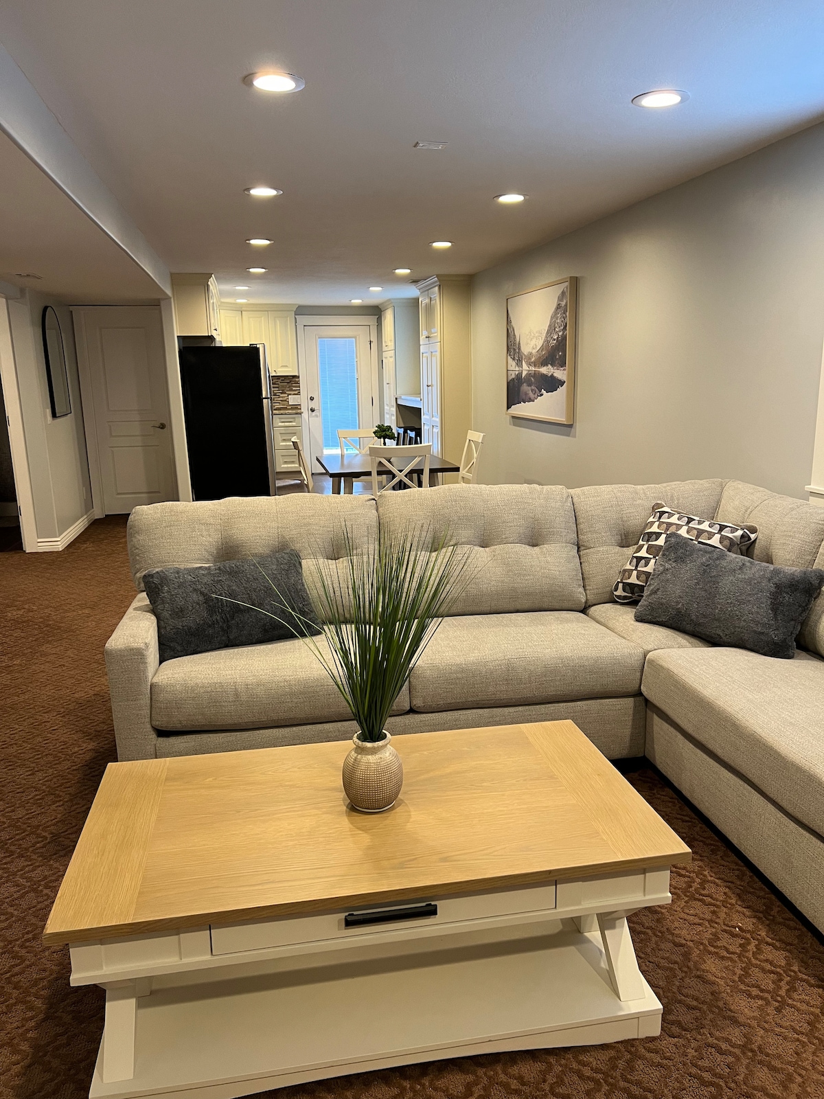 A modern living area features a light-colored sectional sofa adorned with decorative pillows. A small wooden coffee table, topped with a neutral vase and greenery, sits in front. The open layout includes glimpses of a dining area and kitchen in the background.