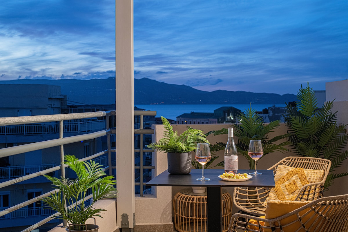 A cozy balcony setting is featured, complete with a small table and two glasses of wine, accompanied by a bottle and snacks. Green plants provide a touch of nature, while a serene view of the water and mountains is visible in the distance under a dusk sky.