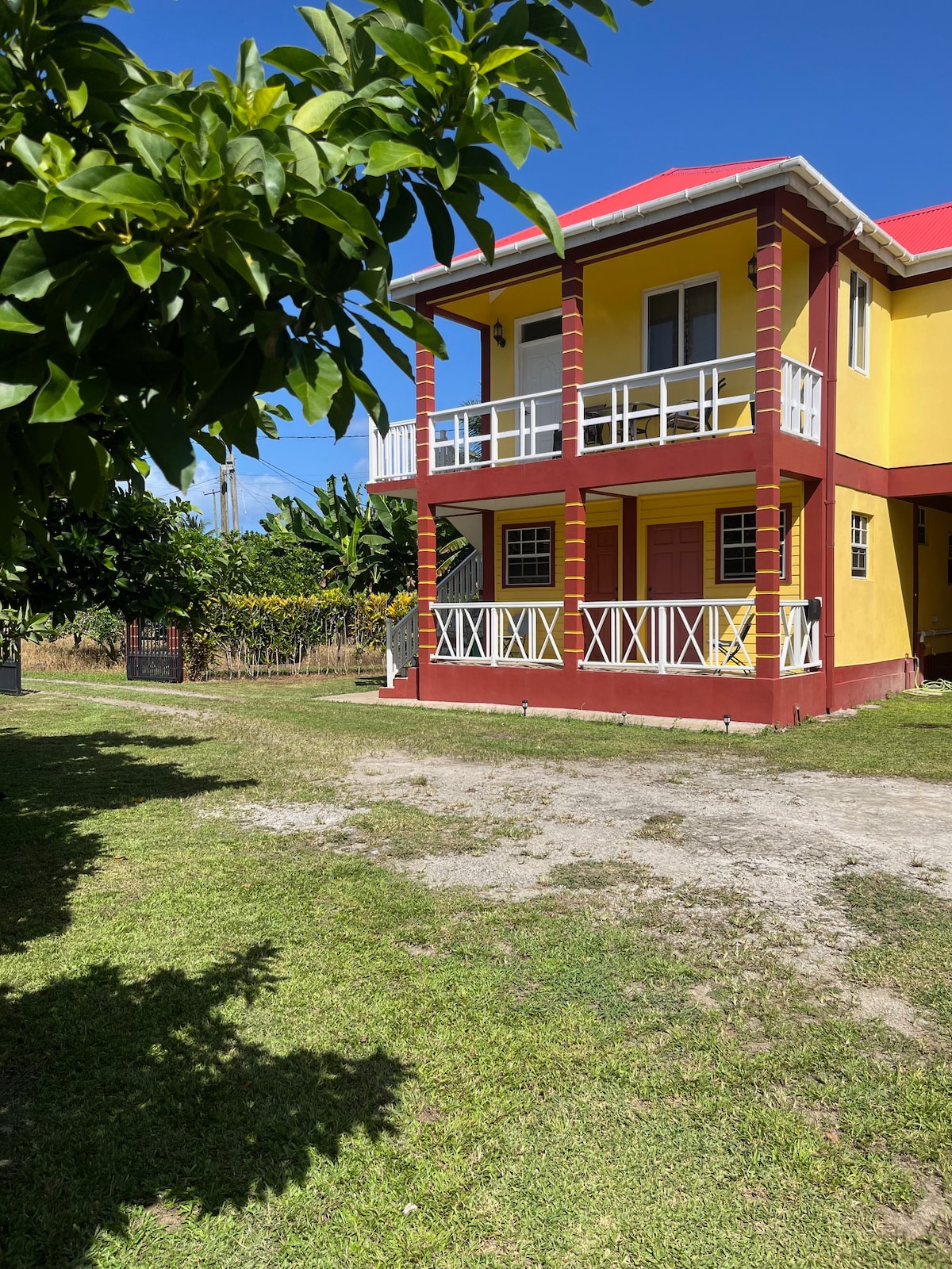 A two-story yellow building is shown, featuring a red roof and white railings on the balconies. Lush greenery surrounds the property, with a grass lawn leading to the entrance. Bright sunlight illuminates the scene, enhancing the inviting appearance of the home.