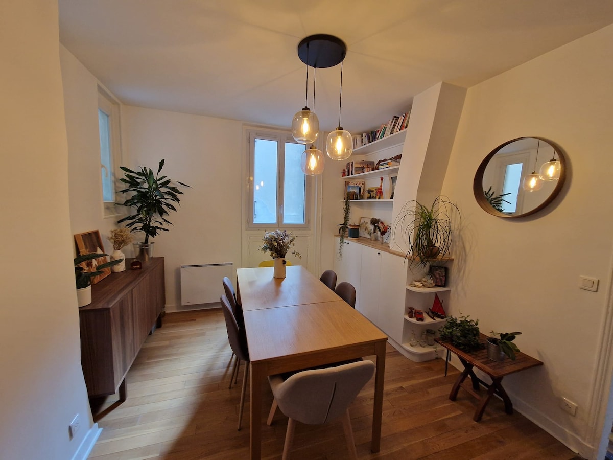 A dining area features a wooden table surrounded by multiple chairs. A warm light fixture hangs above, providing illumination. Shelves line the wall, showcasing books and decorative items. A potted plant adds a touch of greenery, while natural light filters through nearby windows.