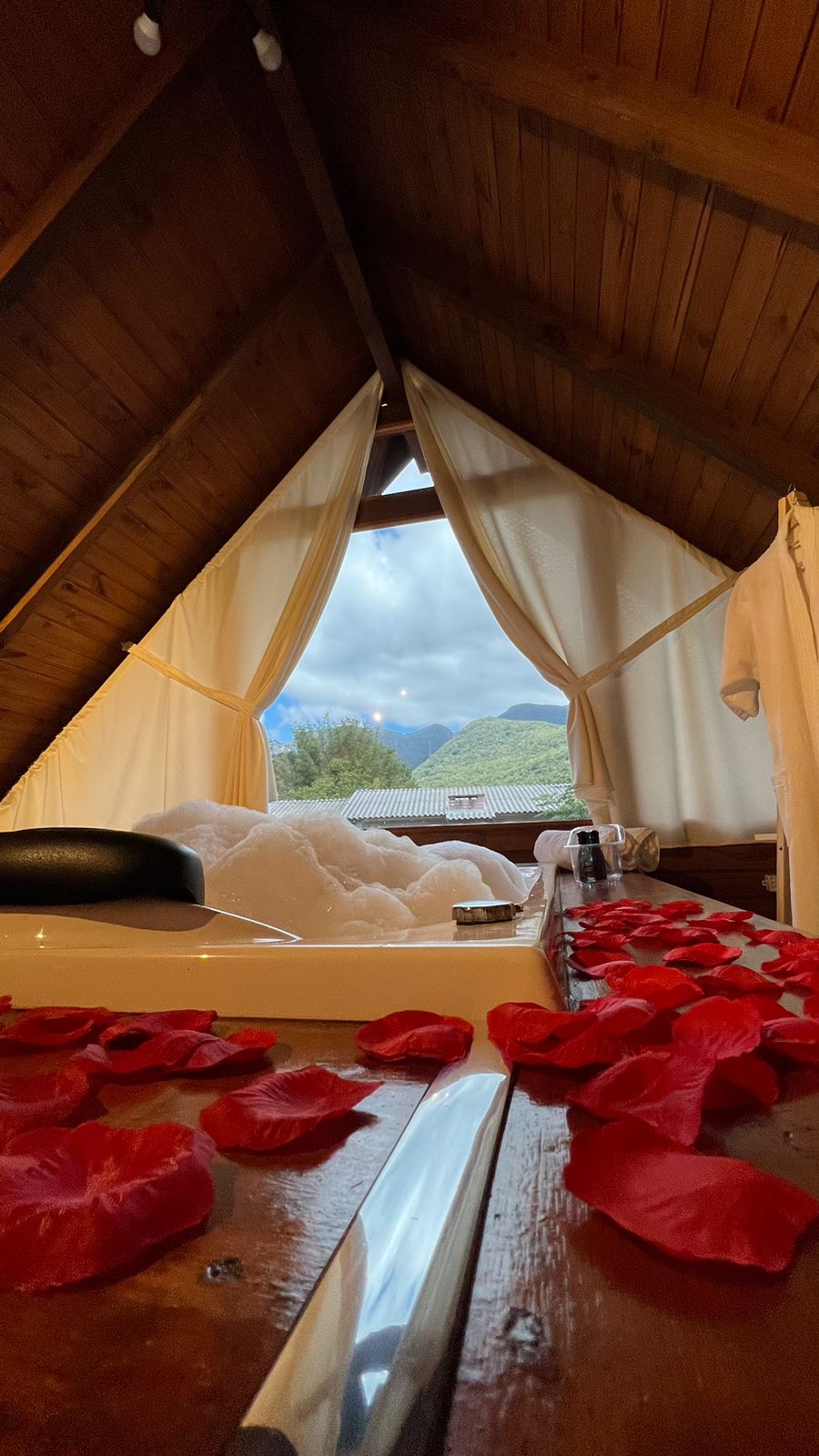A cozy corner features a hydromassage bathtub adorned with rose petals. Soft white curtains frame a view of the surrounding hills, while warm wooden beams add to the tranquil ambiance. Light filters through the space, creating a relaxed atmosphere.