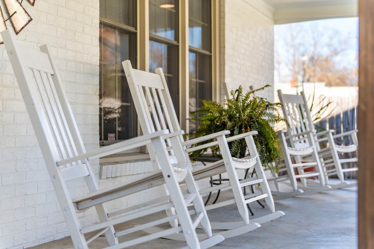 A charming rocking chair front porch features four white rocking chairs positioned along a brick wall. Lush greenery from potted ferns enhances the inviting outlook. Large windows allow natural light to fill the area, creating a peaceful setting for relaxation.