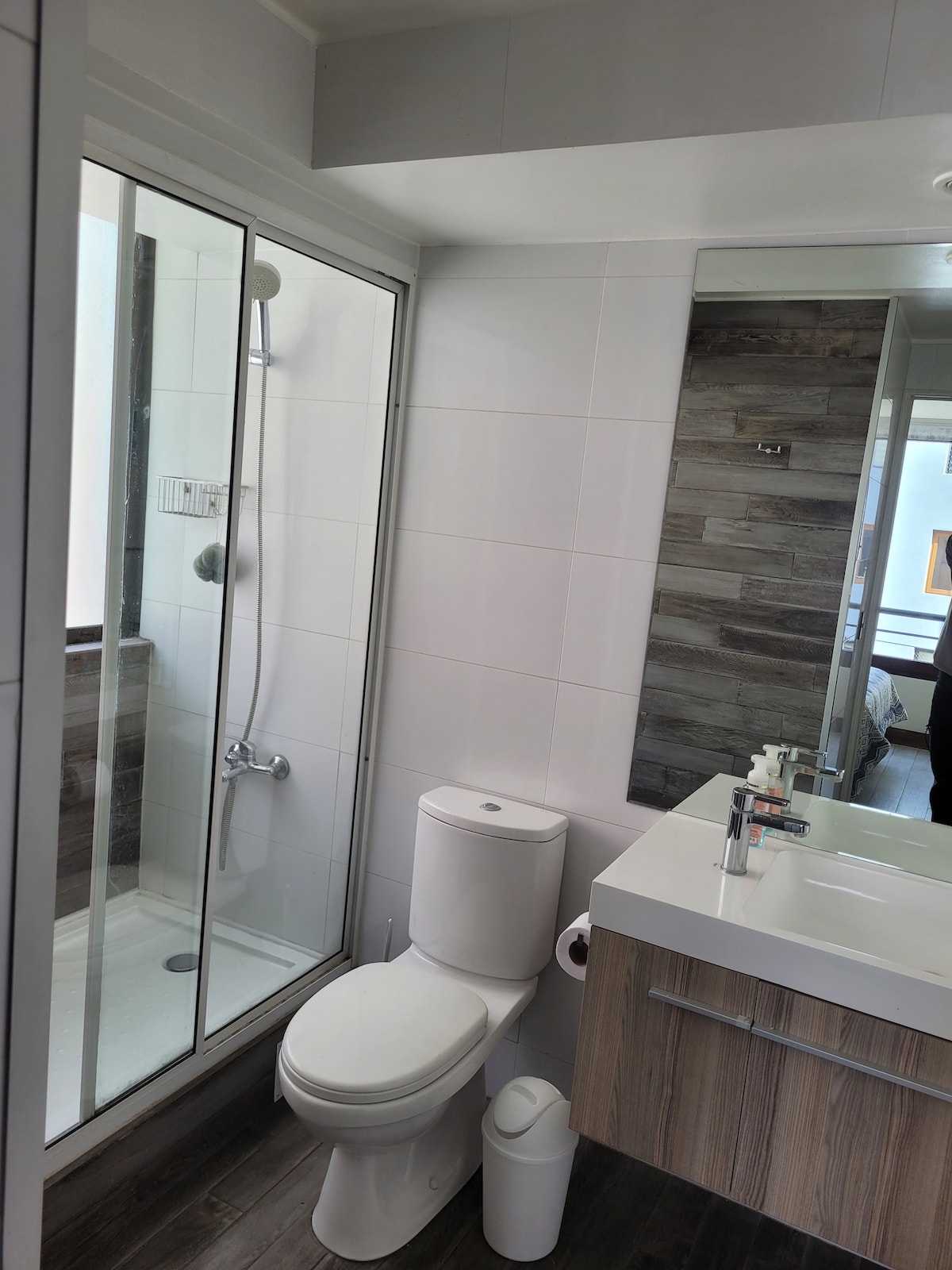The modern bathroom features a glass-enclosed shower with a rainfall showerhead. A sleek vanity with a white countertop and wooden cabinetry is positioned beside a full-length mirror. A clean and minimalist design is highlighted by light-colored tiles and wooden accents.