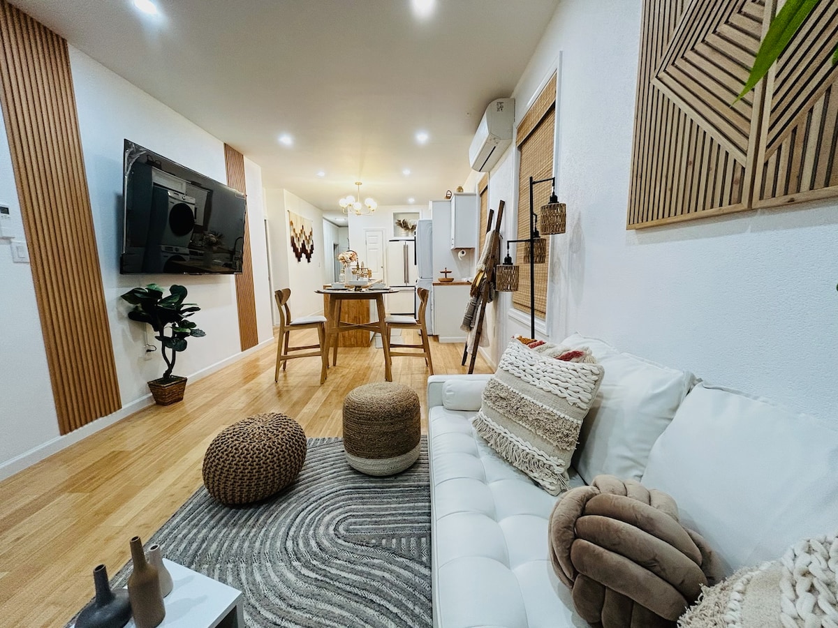 A modern living space features a white sofa adorned with textured cushions and a circular area rug. A dining area with wooden chairs is visible in the background, accompanied by decorative wall art and a potted plant that adds a touch of greenery.