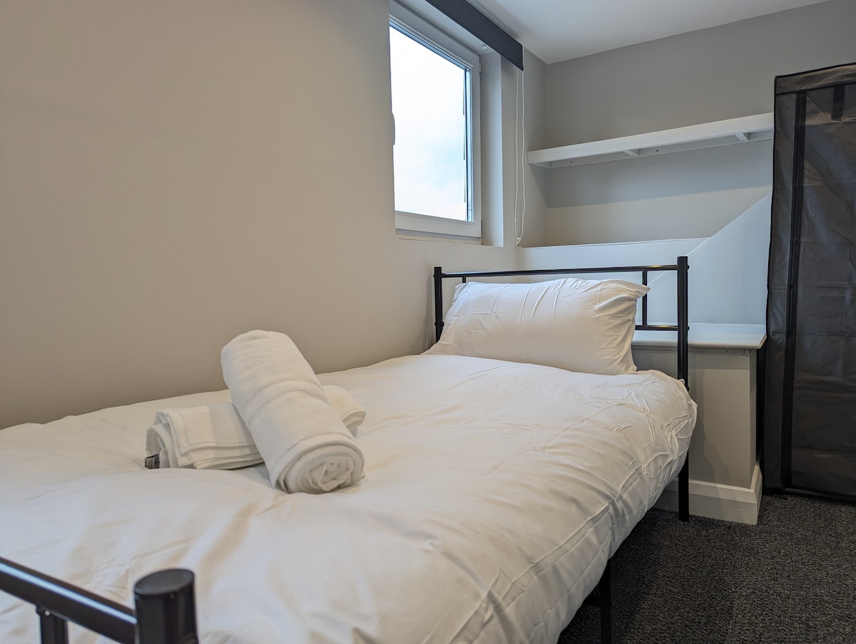 A single bed is set against a light grey wall, featuring fresh white linens and rolled towels. A nearby window allows natural light to enter, illuminating a minimalist workspace with a shelf above. A closet is positioned next to the bed, enhancing storage options.