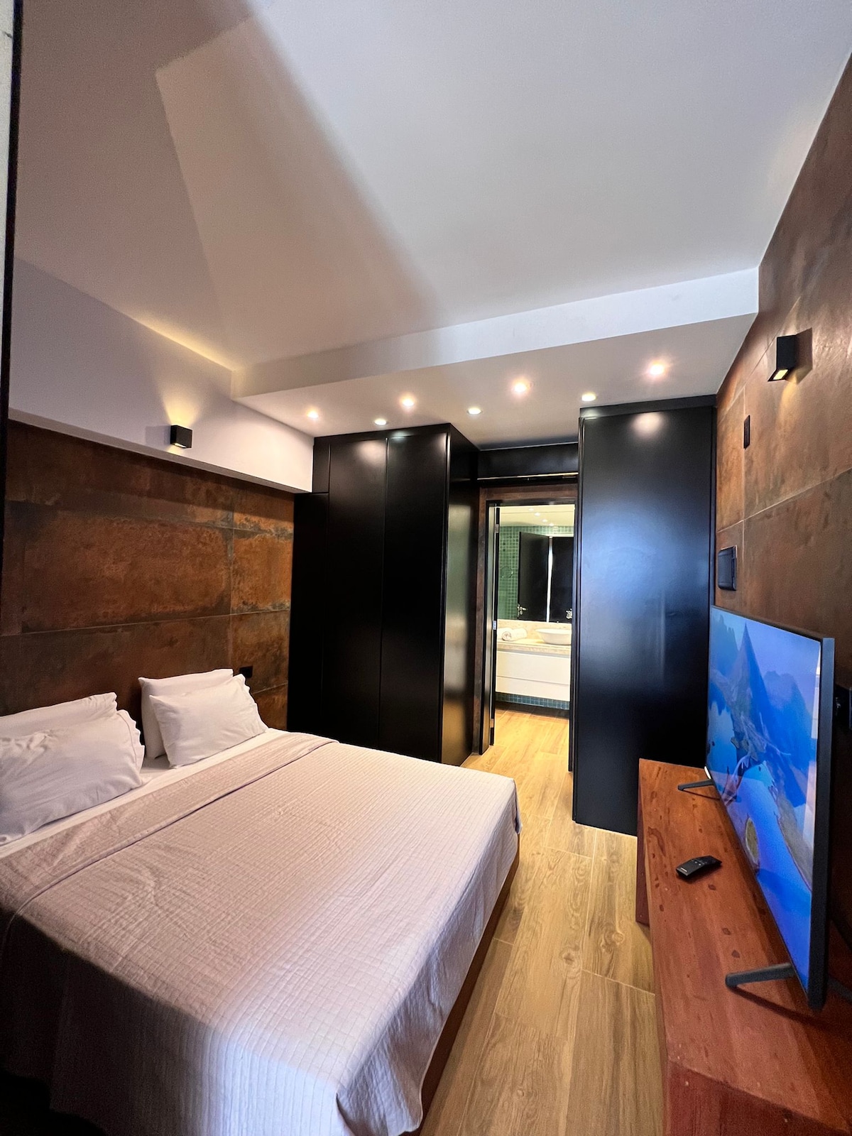 A modern studio bedroom features a spacious bed with crisp linens. The walls are adorned with sleek dark panels, and recessed lighting provides soft illumination. A TV is positioned on a wooden console, while a glass door leads to a visible bathroom area.