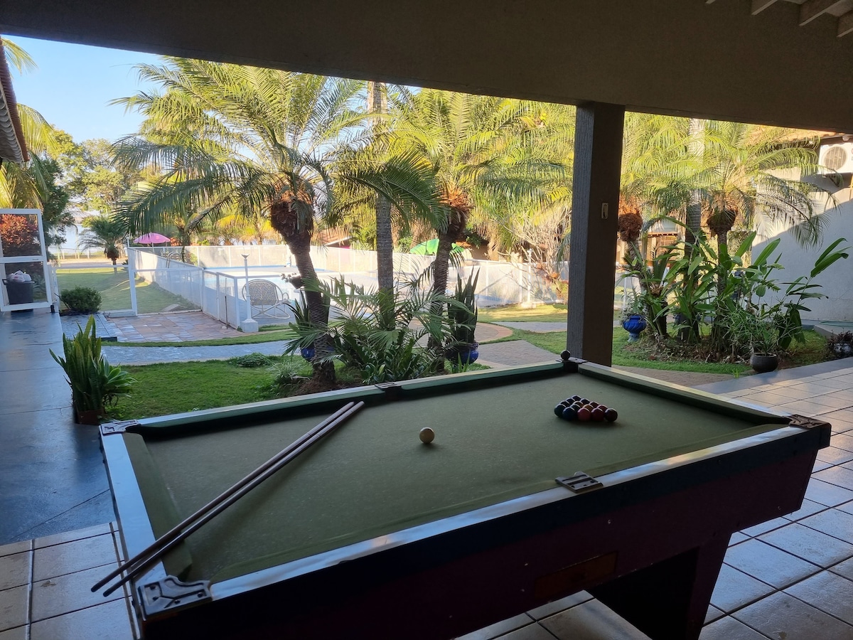 A billiard table with cues and balls is positioned in a covered area. Through large openings, a green outdoor space is visible, featuring palm trees and a swimming pool in the background. The setting offers a relaxed atmosphere for leisure activities.