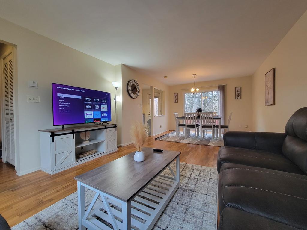 The living room features a dark leather couch facing a large flat-screen TV mounted on the wall. A light wooden coffee table sits on a soft area rug. Natural light enters through multiple windows, illuminating the space, with a dining area visible in the background.