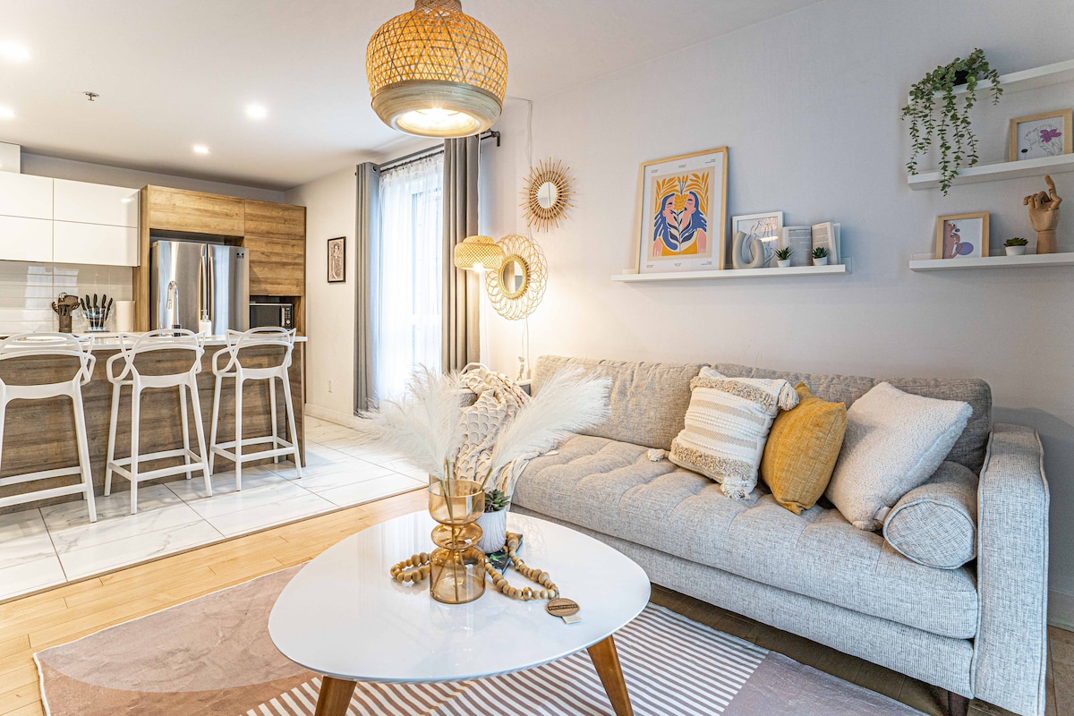 A modern living area features a comfortable gray sofa accompanied by decorative pillows. A round coffee table displays a vase with pampas grass and decorative items. The backdrop includes a well-equipped kitchen with wooden cabinetry and high stools, with natural light streaming through sliding glass doors.