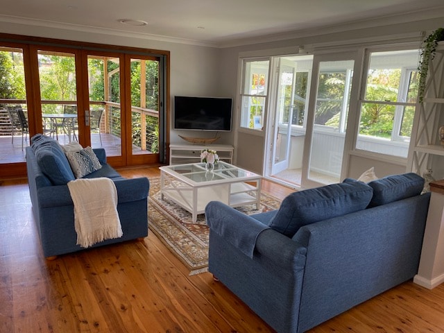 A comfortable living area features two blue sofas facing a white coffee table on a wooden floor. Large glass doors open to a deck, allowing natural light to fill the space. A television is positioned near the window, adding functionality to the room.