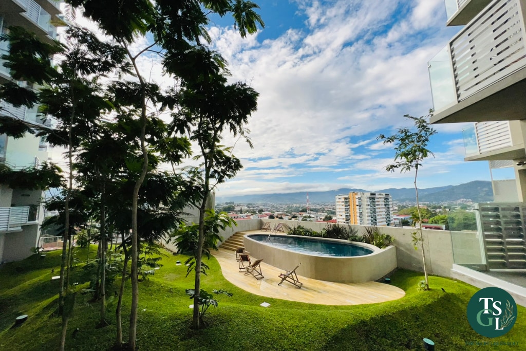 A serene outdoor area is presented, featuring a circular pool surrounded by lush greenery and wooden deck chairs. The view encompasses urban buildings and distant mountains under a bright sky, enhancing the tranquil setting.