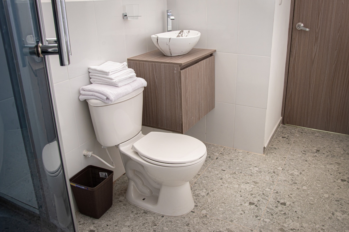 A clean bathroom features a modern design with a wall-mounted sink atop a wooden cabinet. Fresh towels are neatly stacked beside the toilet, accompanied by a small wastebasket. The light-colored tiled walls complement the textured flooring.