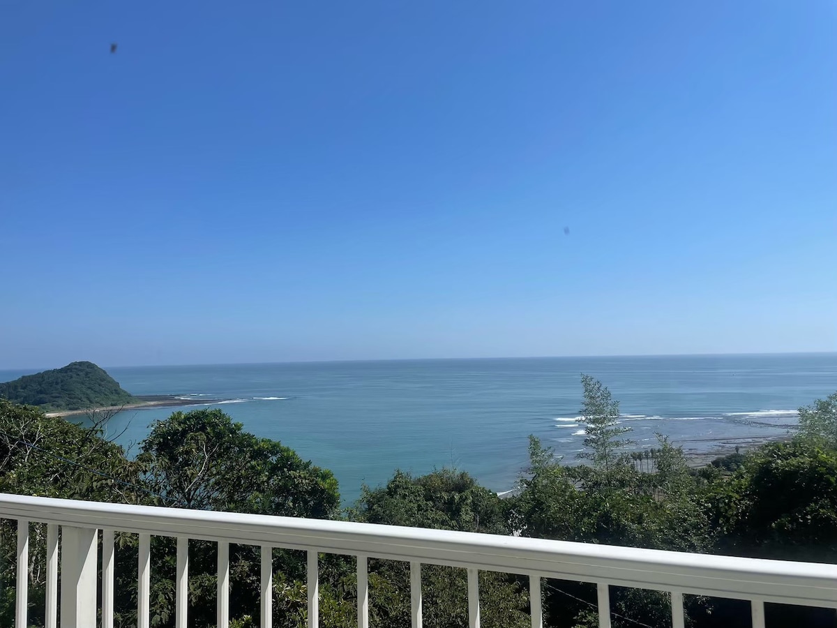 An expansive view of the Pacific Ocean is presented from a balcony. The serene blue waters stretch to the horizon, framed by lush greenery in the foreground. Clear skies enhance the tranquil atmosphere of the coastal landscape.