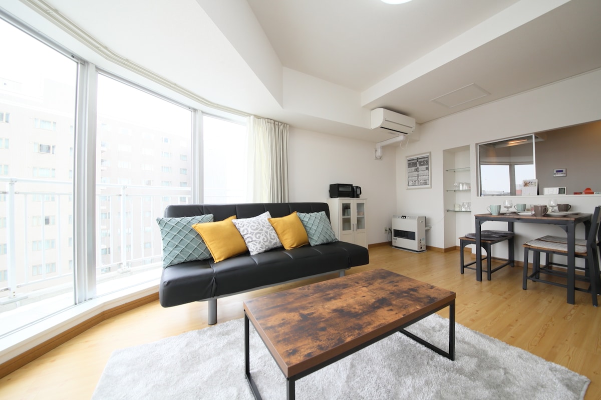 A bright living area features a modern black sofa adorned with yellow and patterned pillows. A wooden coffee table rests on a soft rug, complemented by an open dining space in the background. Large windows allow natural light to fill the room.