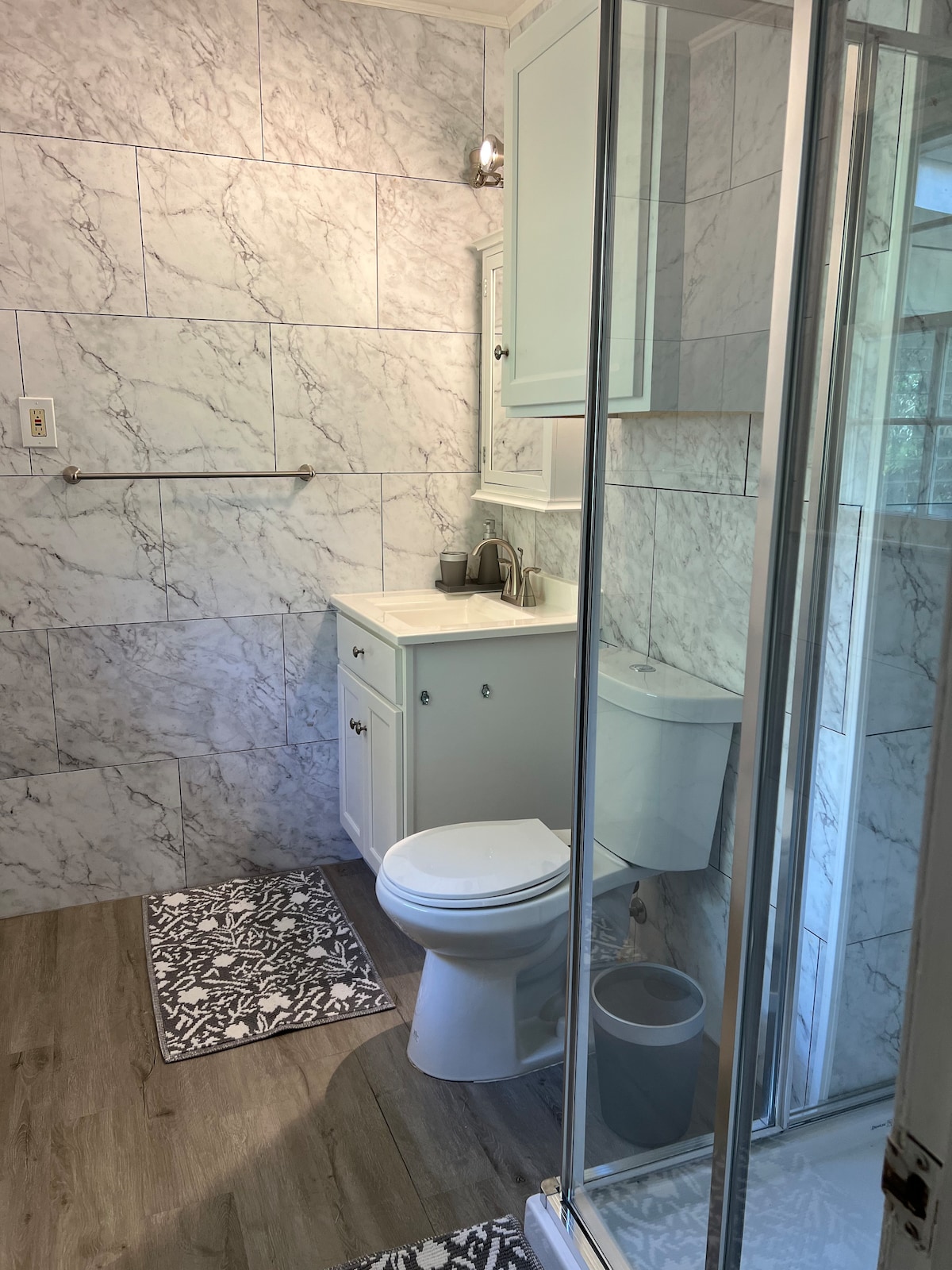A modern bathroom is showcased with marble-patterned walls and a spacious shower enclosure. A white vanity is equipped with a sink and storage, while gray accent rugs lie on the floor. Natural light filters in, enhancing the room's clean aesthetics.