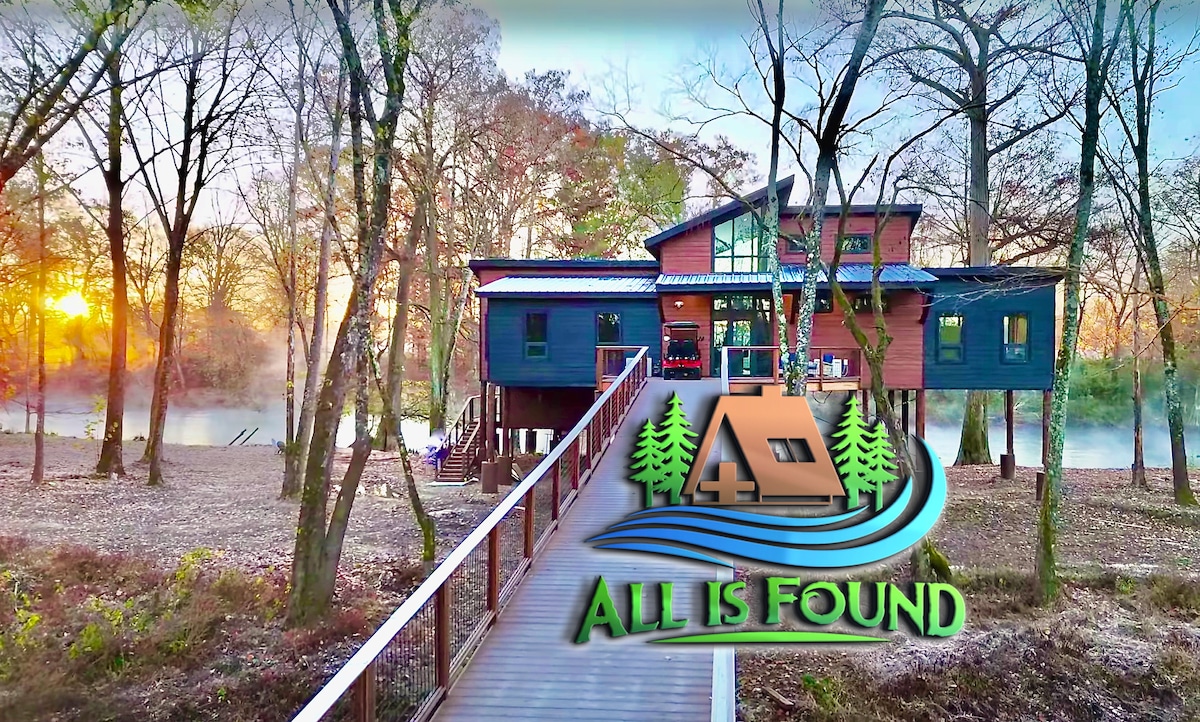 A contemporary cabin is situated along the riverbank, featuring a multi-level design and large windows. A wooden bridge leads to the entrance, surrounded by trees. The warm hues of sunset illuminate the scene, reflecting on the river's surface.
