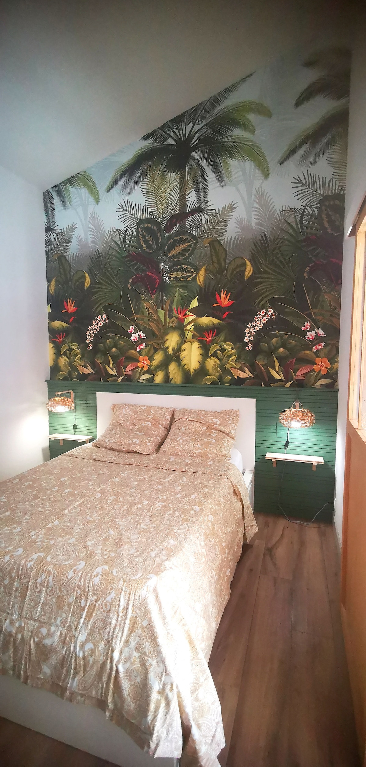 A cozy sleeping area features a spacious bed adorned with a neutral-toned patterned duvet. A vibrant tropical mural covers the wall behind, depicting lush greenery and colorful plants. Subtle bedside lights are positioned on either side, illuminating the serene space.