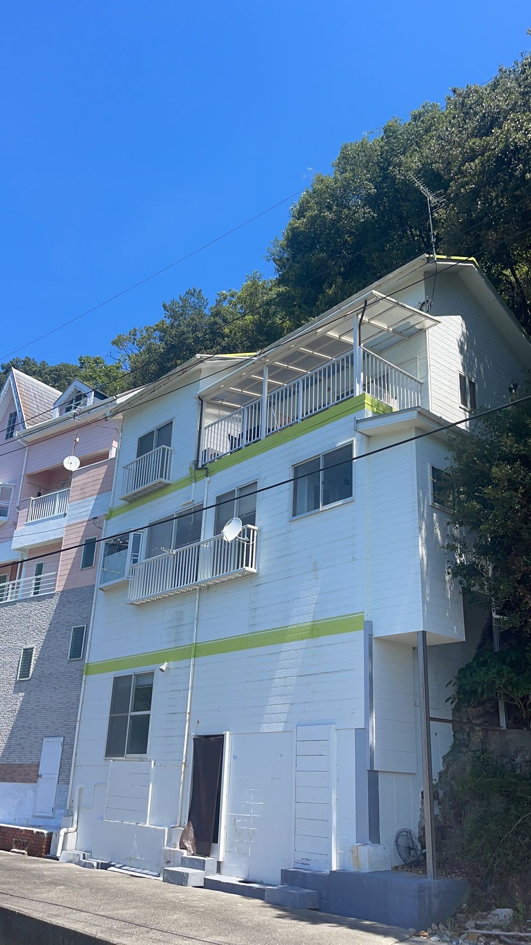 The three-story villa features a white exterior with green accents and multiple balconies. Satellite dishes are mounted on the walls, and the surrounding hillside is lush with greenery. A driveway leads to the entrance, providing easy access to the property.