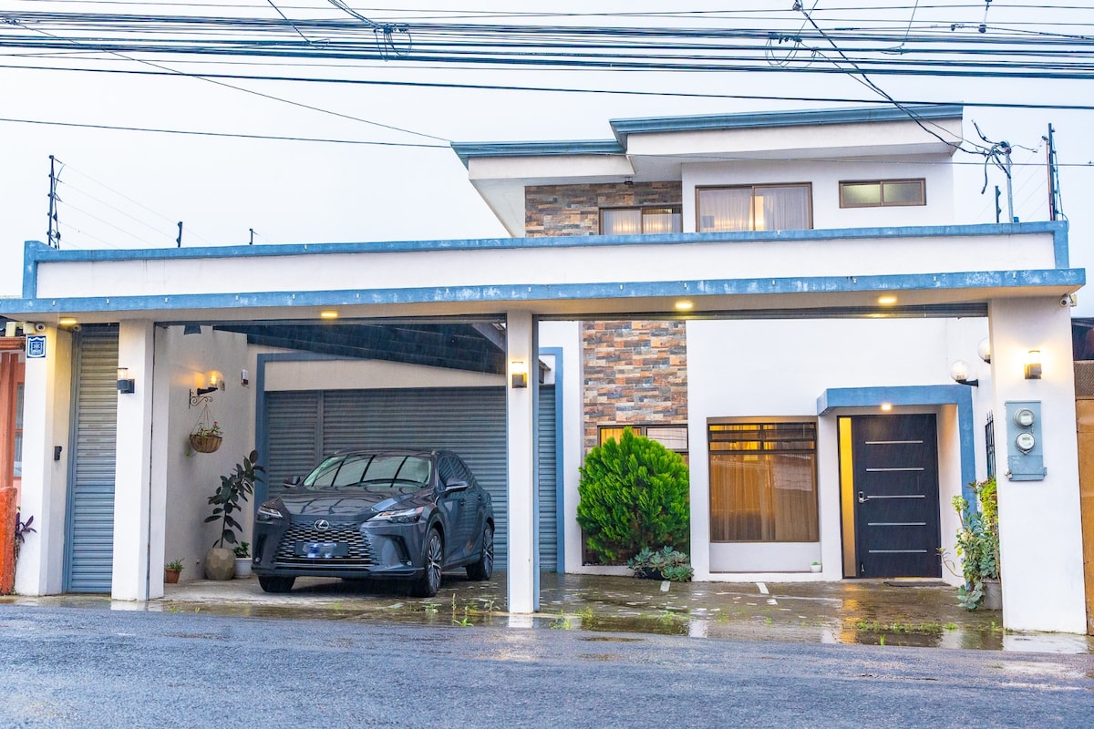 A modern two-story house is featured, with a spacious driveway accommodating a vehicle. The entrance is marked by a solid door, and large windows allow natural light to enter. The exterior displays a blend of stone and stucco finishes, enhancing its architectural appeal.