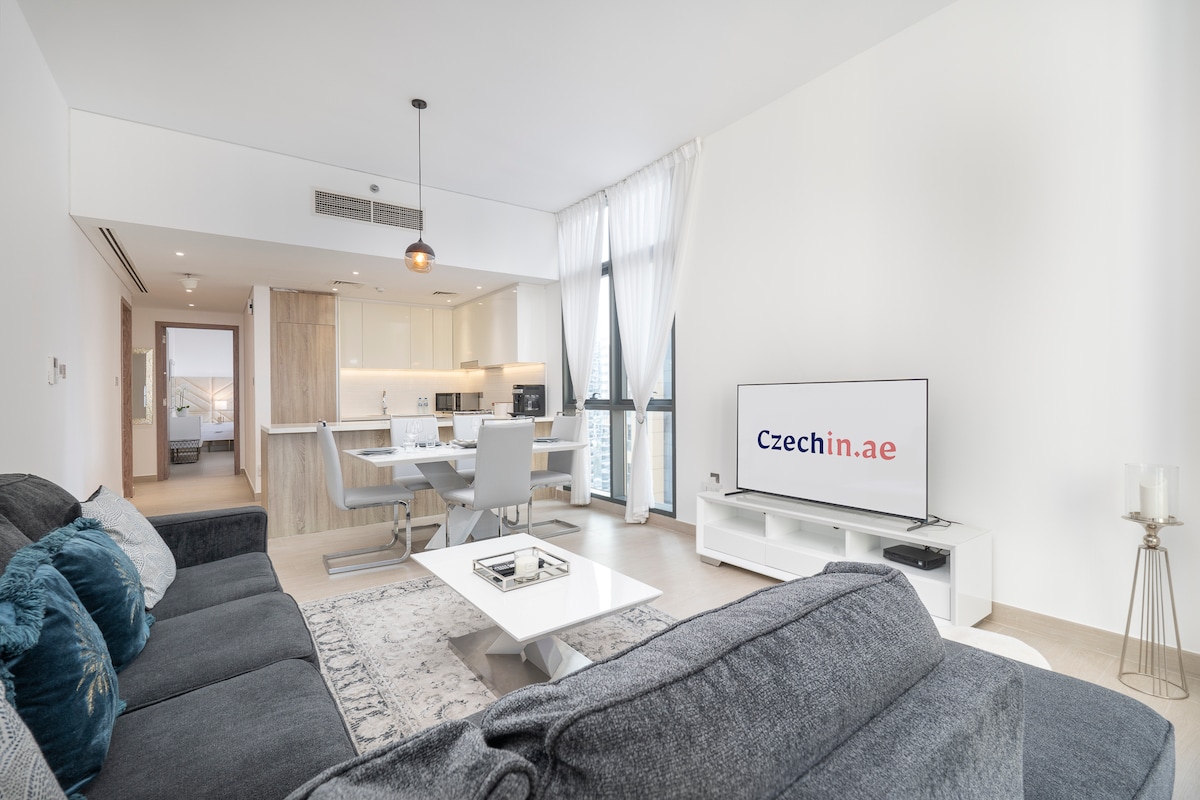 The living area features a comfortable dark grey sofa, a coffee table, and an entertainment unit with a television. A dining table with white chairs is visible in the background. Large windows allow natural light to fill the space, with sheer curtains adding a soft touch.