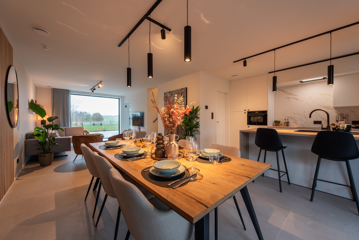 A well-lit dining area features a wooden table set for a meal, surrounded by comfortable chairs. An open-plan layout connects to a living space with a cozy seating arrangement. Large windows provide views of the greenery outside, with modern lighting fixtures enhancing the ambiance.