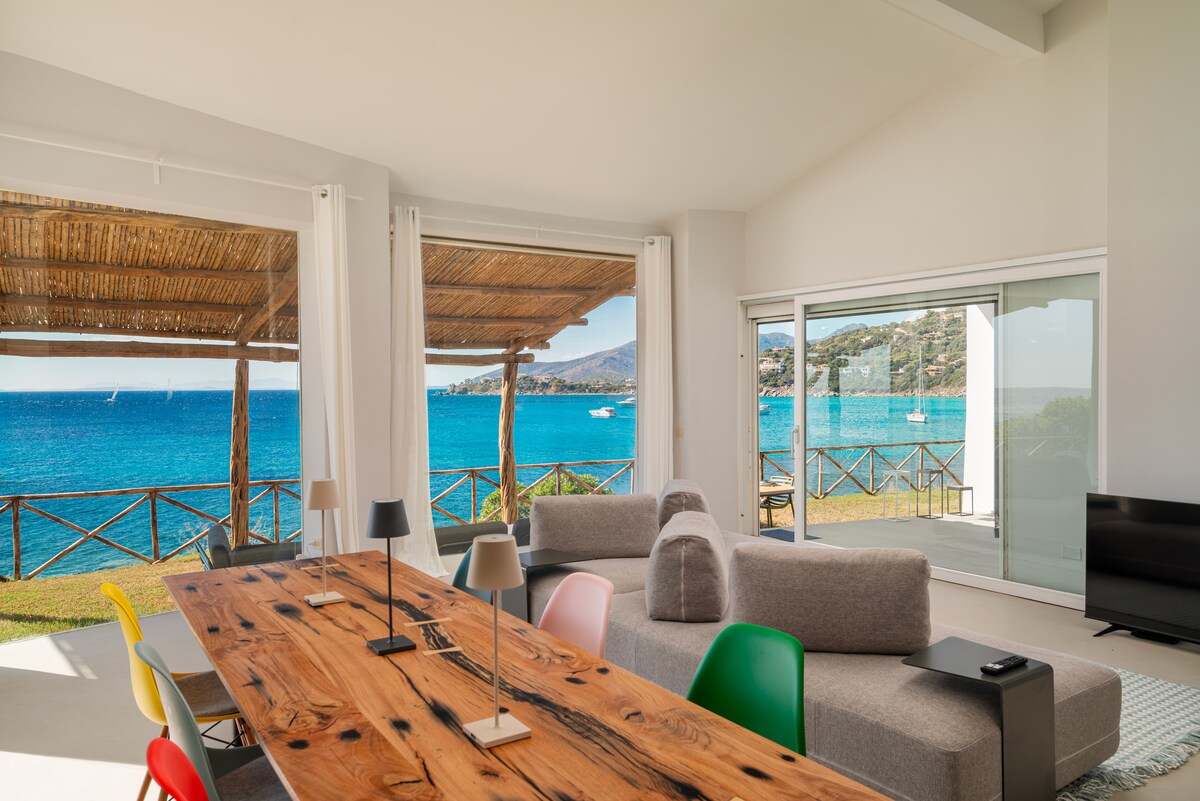 A modern living and dining area features large glass windows that provide uninterrupted views of the sea. The spacious table made of natural wood is surrounded by colorful chairs. The room is illuminated by natural light, highlighting the minimalist décor.
