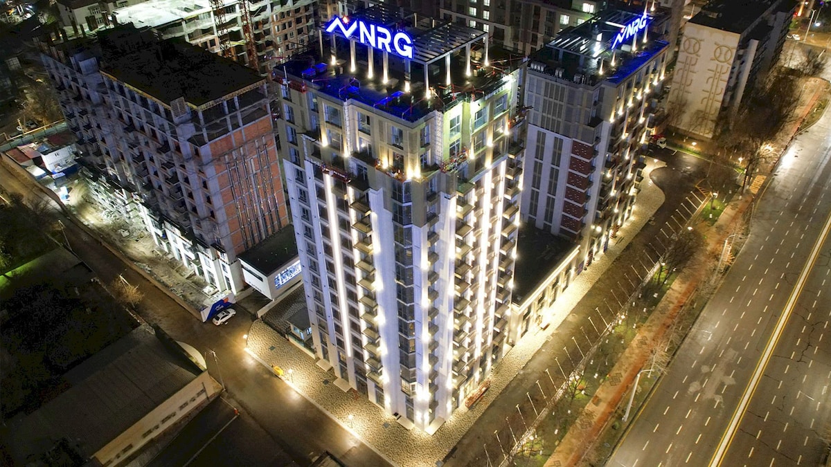 An aerial view of a modern residential building illuminated at night. The structure features multiple stories, balconies with lights, and a prominent logo on the rooftop. Surrounding streets and low-rise buildings are visible, enhancing the urban setting.