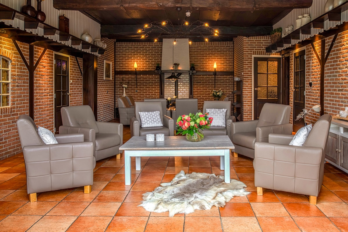 A spacious living area features six comfortable armchairs arranged around a wooden coffee table. Soft lighting is provided by decorative fixtures above, while a textured area rug adds warmth to the tiled floor. Exposed brick walls contribute to a cozy and inviting ambiance.
