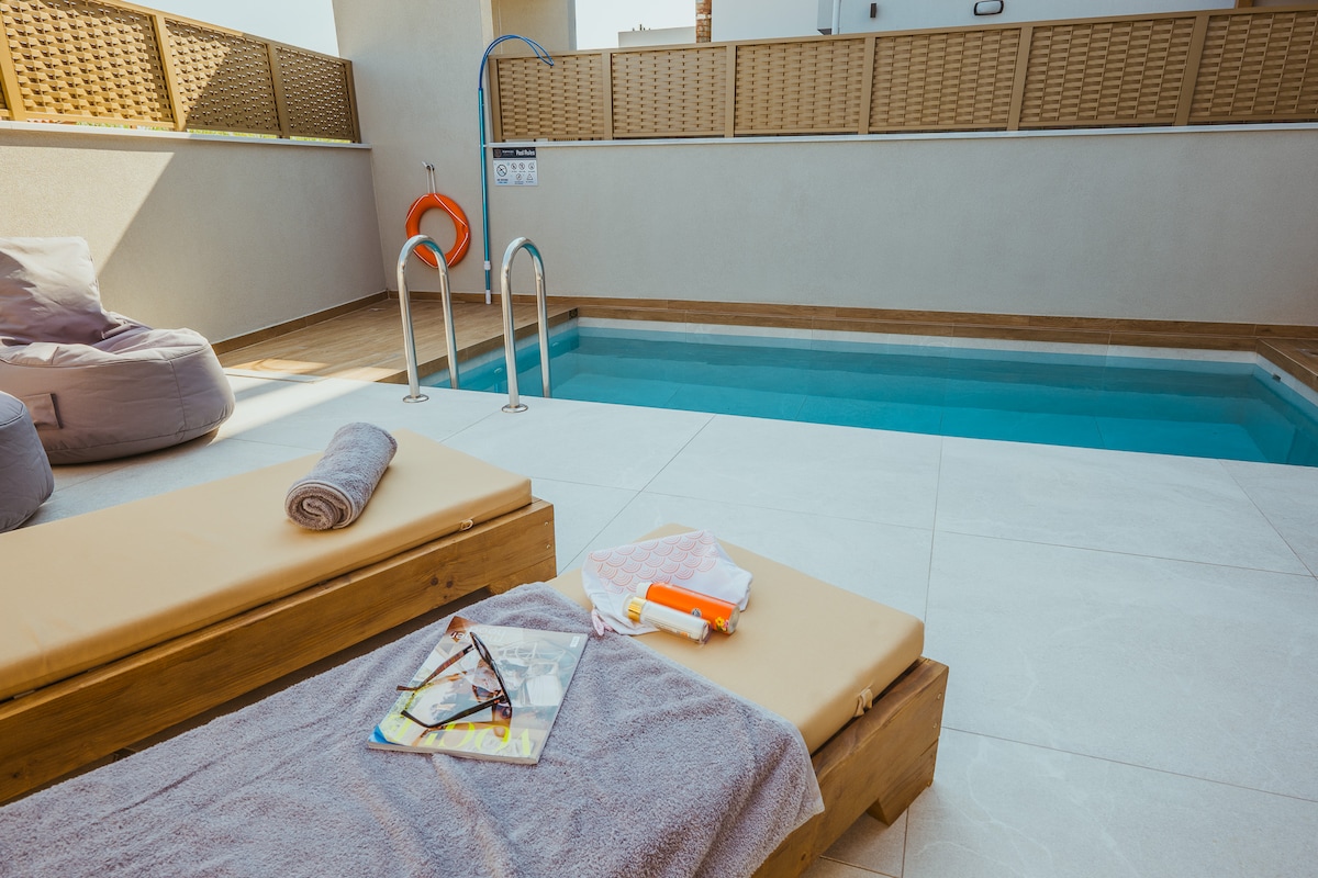 A private pool area is displayed, featuring a clear, inviting swimming pool surrounded by light-colored tiles. Wooden lounge beds are positioned nearby, adorned with rolled towels. A pair of sunglasses and magazines rest on the surface, with a lounge area visible in the background.