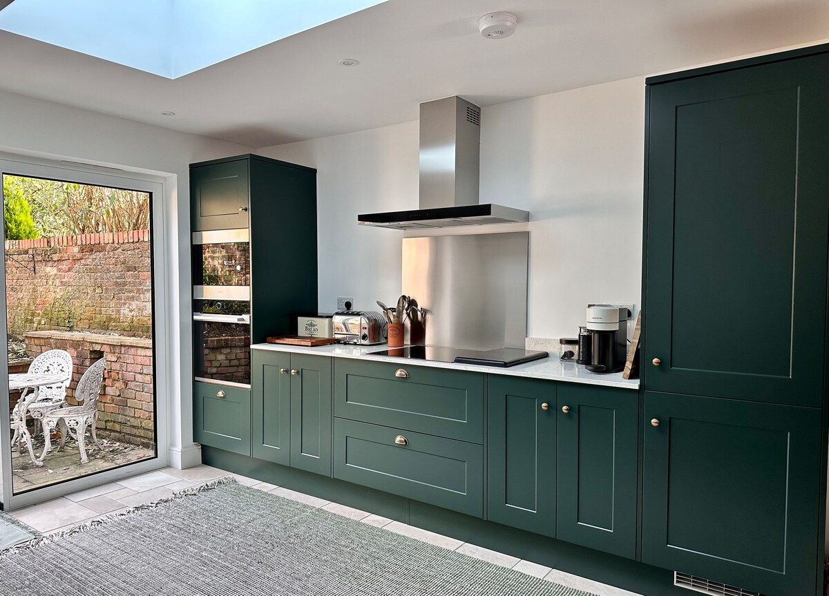 A modern kitchen features deep green cabinetry and a spacious countertop that showcases stainless steel appliances. A large window offers views of the private garden, and a cozy dining set is visible outside through the glass door.