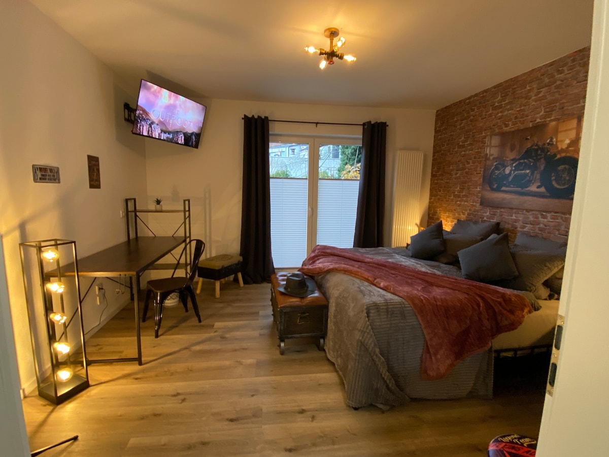 A cozy room features a spacious bed adorned with multiple pillows and a soft blanket. A large window allows natural light, partially covered by dark curtains. A wall-mounted TV is visible above the bed, while a desk and chair are positioned nearby, enhancing the functionality of the space.
