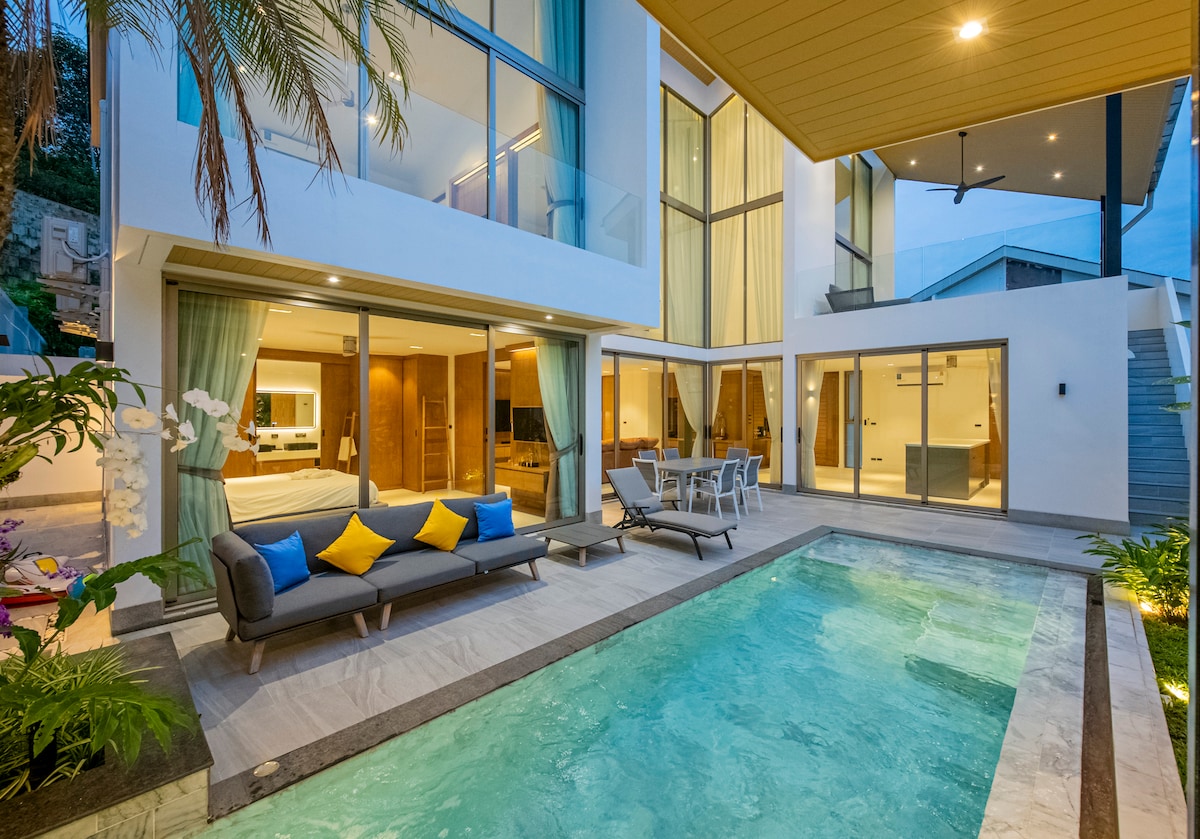 The outdoor area features a private pool surrounded by landscaped greenery. Comfortable lounge seating with colorful cushions is positioned nearby, while large windows of the villa reflect the inviting atmosphere. The integration of indoor and outdoor spaces is evident, enhancing accessibility and enjoyment.