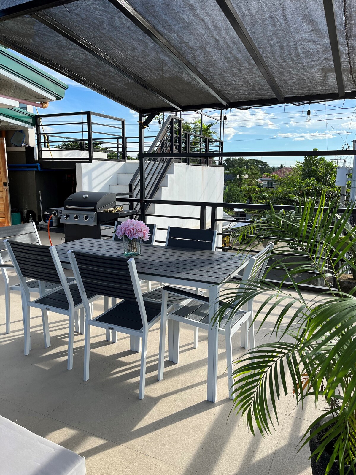 An outdoor dining area features a gray table surrounded by six chairs, set against a backdrop of greenery and a distant staircase. A grill is positioned to one side. Sunlight filters through a canopy overhead, creating a shaded but bright space for gatherings.
