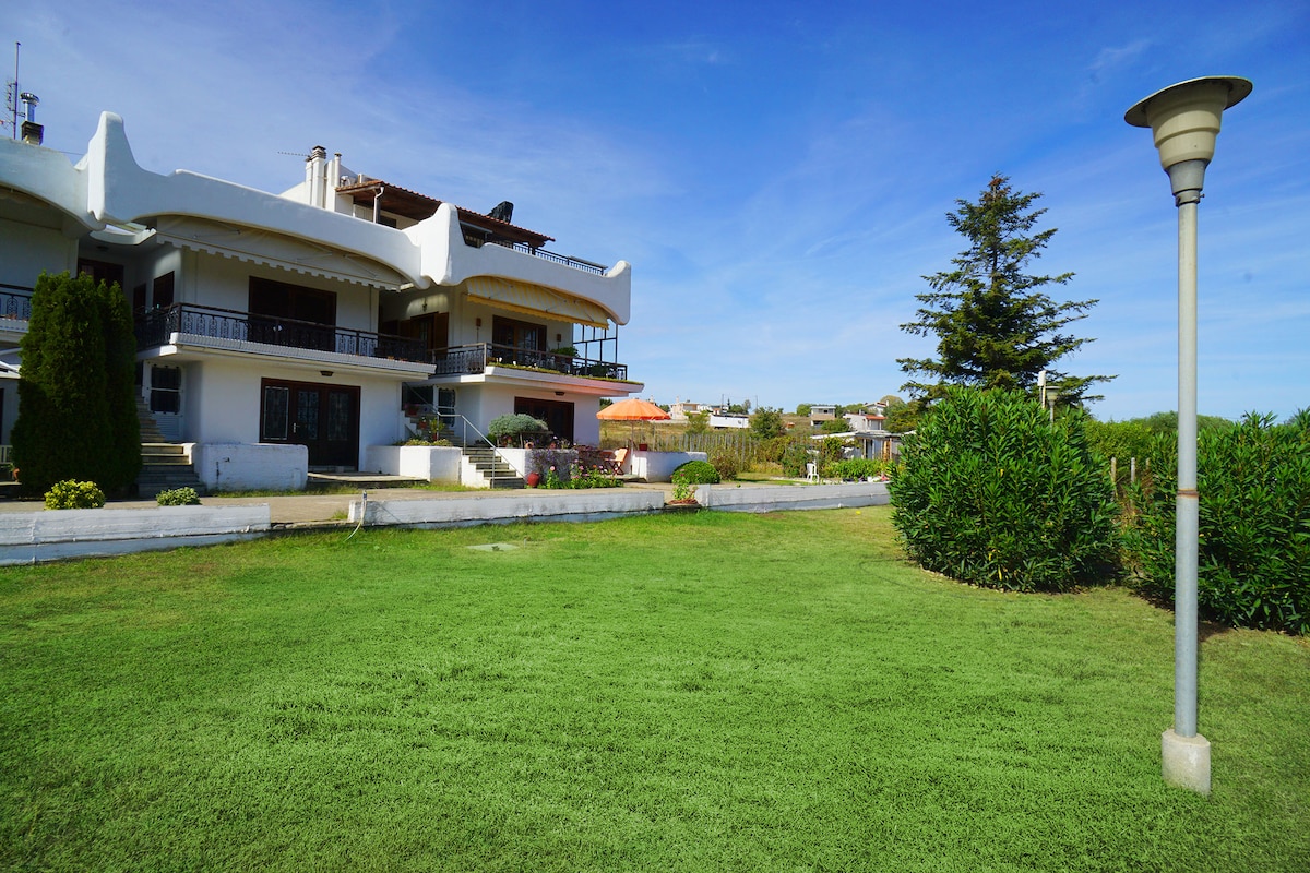 The exterior view showcases a three-story villa with distinct architectural features. Lush green grass surrounds the building, with well-maintained shrubs and trees adding to the landscape. A lamp post stands in the foreground, contributing to the inviting outdoor environment.