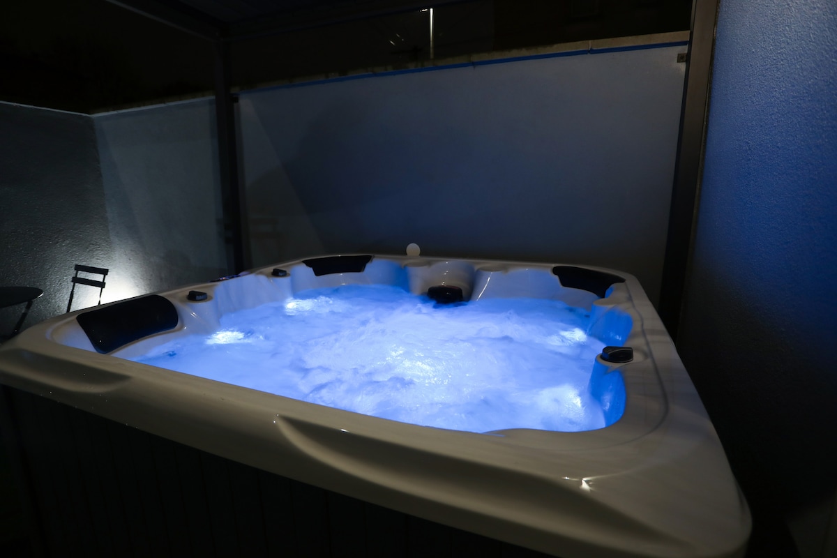 An outdoor spa is located within a private enclosure. The spa features a soothing blue illumination, creating a tranquil ambiance. Soft bubbles emerge from the jets, inviting relaxation. The surrounding area is enclosed with walls, providing privacy and a serene atmosphere.