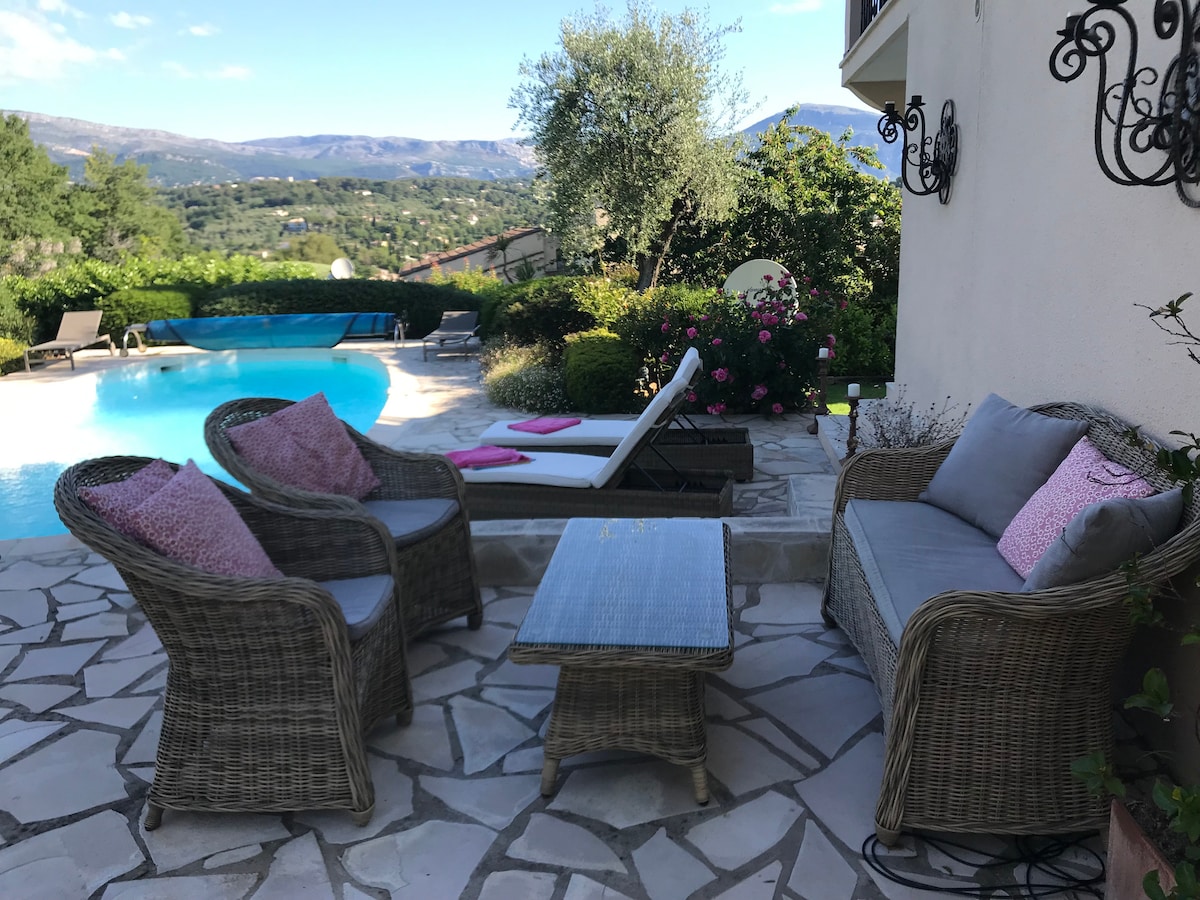 An outdoor seating area features woven chairs with soft cushions and a low table, set on a stone patio. In the background, a tranquil pool reflects the surroundings, with lounge chairs positioned nearby. Lush greenery and blooming flowers provide a serene backdrop.