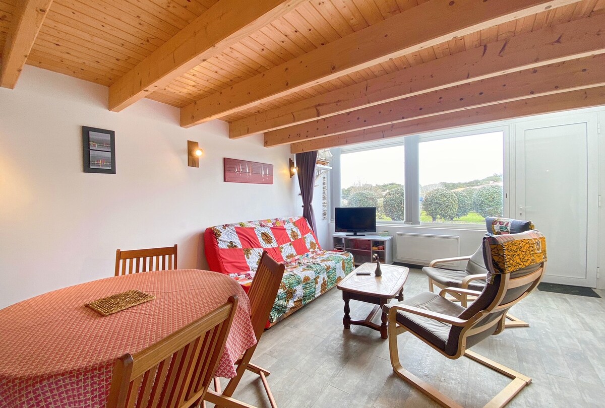 A bright living area features wooden beams overhead and a large window offering views of the outside greenery. A sofa bed is adorned with a colorful quilt, accompanied by a round dining table and several comfortable seating options, including a lounge chair.