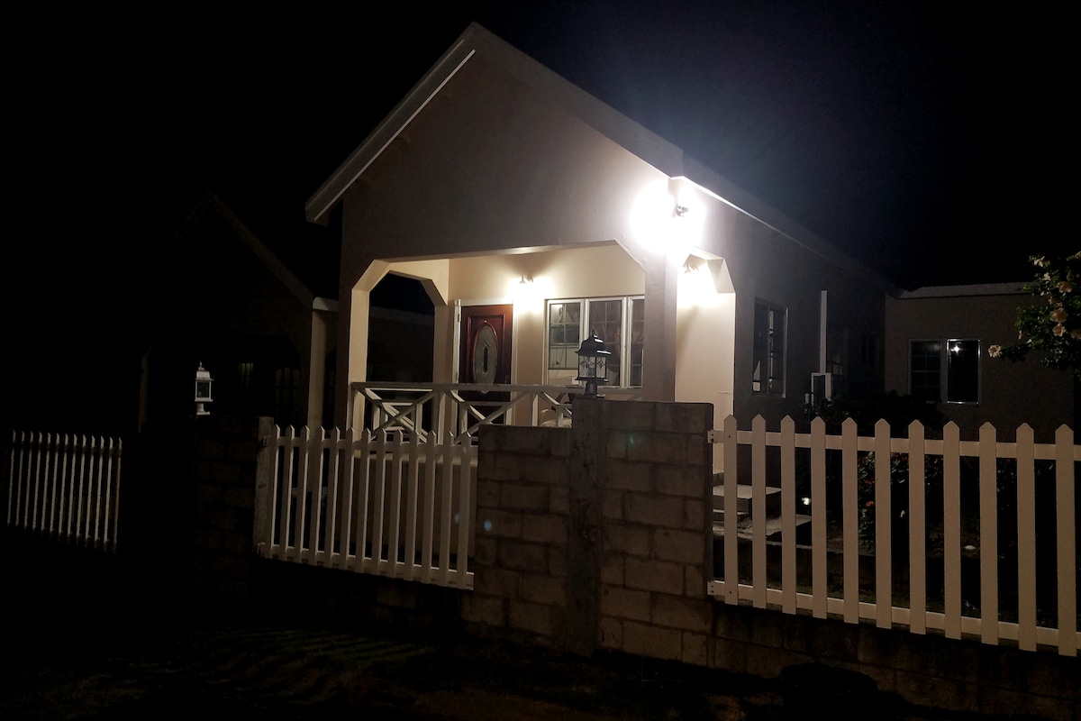 The exterior of a single-story house is shown at night, featuring a softly lit entrance with an arched porch. A decorative light illuminates the front door, while a fenced yard with a white picket fence provides a cozy boundary. Windows reflect the surrounding ambient light.