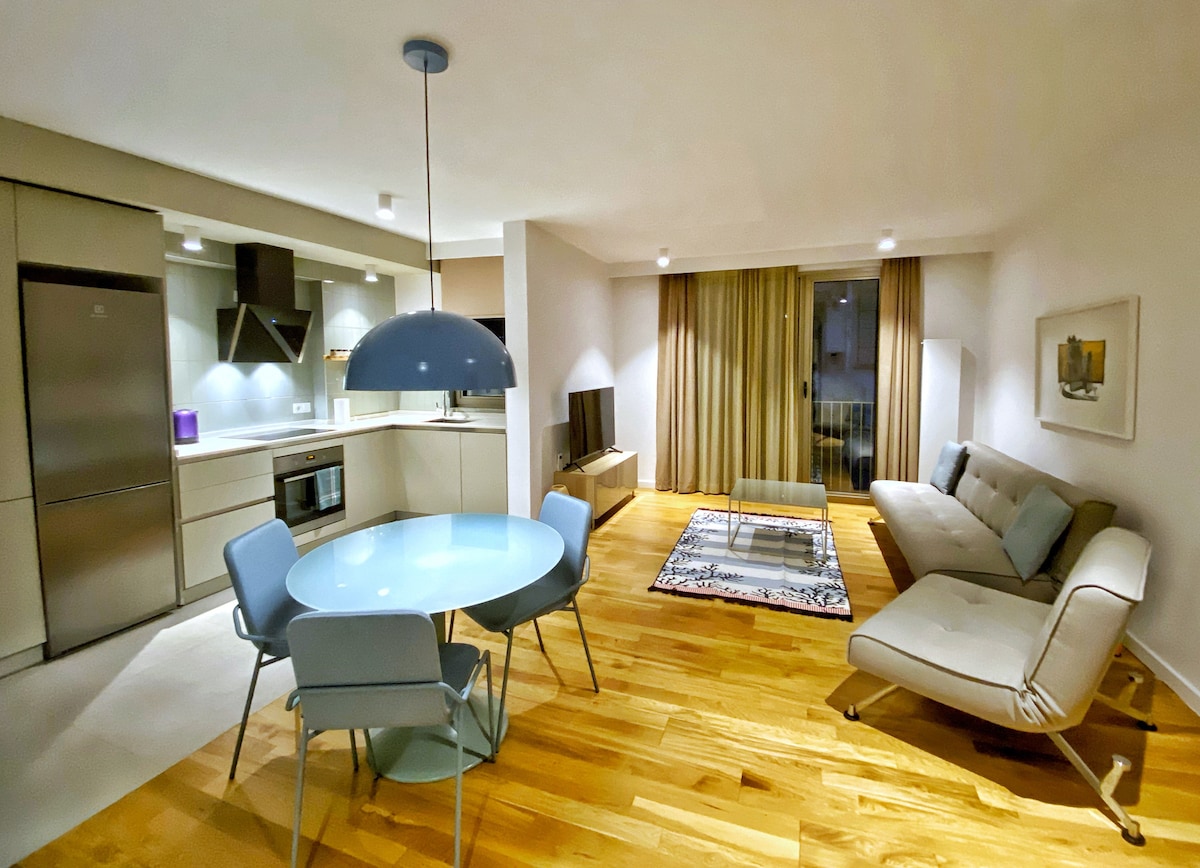 The living area features a light wood floor with a spacious seating area, accented by large windows and soft curtains. A round dining table with four chairs is positioned near a modern kitchen, which includes sleek appliances and a contemporary design.