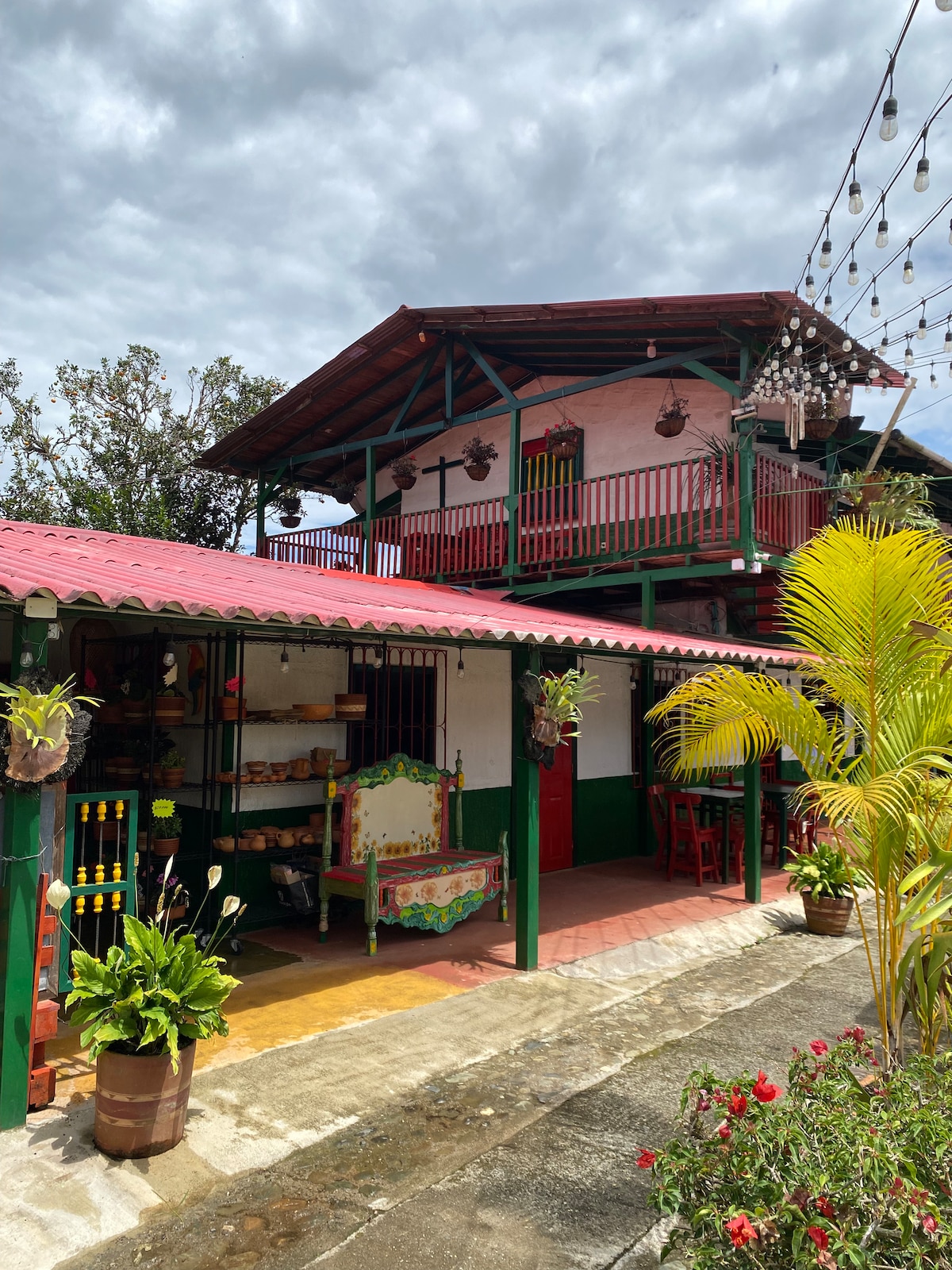 A vibrant house features a two-story design with a red roof and green accents. Intricate balconies are visible on the upper floor, and colorful decorative elements adorn the entrance. Lush plants and flowers enhance the outdoor area, creating an inviting atmosphere.