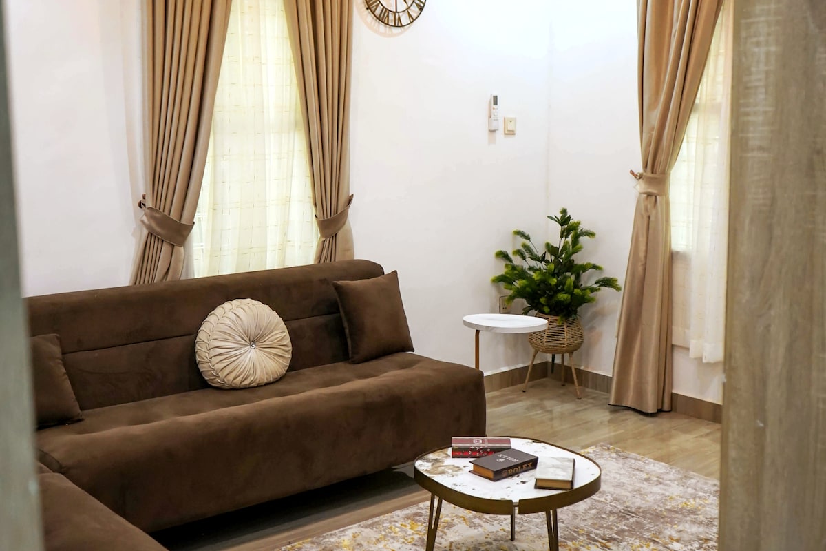 A cozy living area features a deep brown sectional sofa adorned with a round cushion. A small round table, topped with books, is positioned in front of the sofa. Natural light filters through sheer curtains at two windows, enhancing the tranquil ambiance.