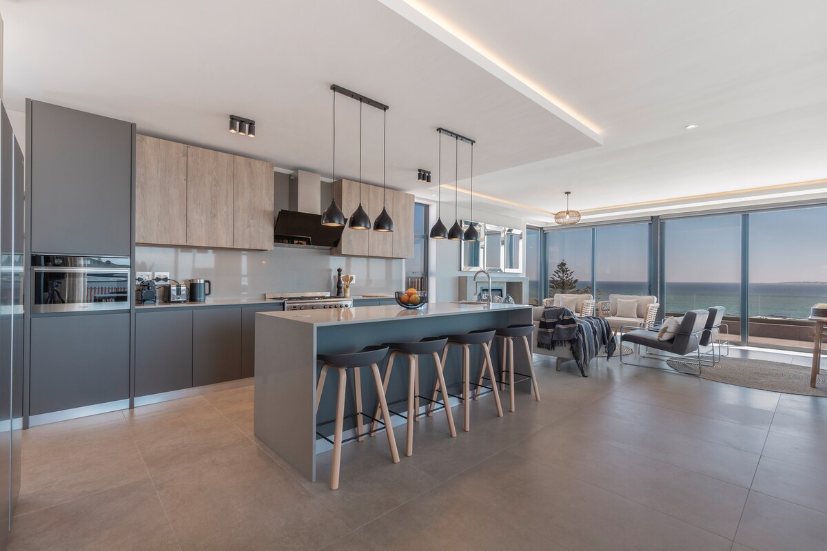 A modern kitchen and living area feature a spacious island with seating for six. Sleek cabinetry in muted tones complements the contemporary design. Large windows provide unobstructed ocean views, allowing natural light to illuminate the open space.