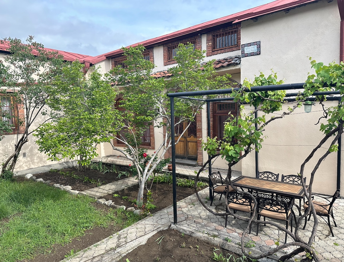 A garden area showcases a stone pathway bordered by green grass and well-maintained trees. A sturdy outdoor dining table surrounded by ornate metal chairs is positioned under a simple pergola. The entrance to the house is visible, featuring traditional wooden doors and windows.