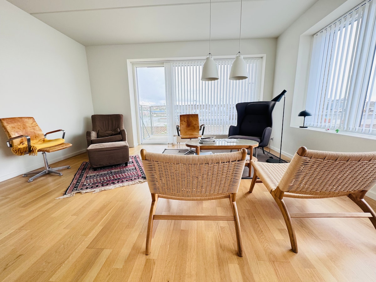 A bright living area is featured with large windows allowing natural light to fill the space. Comfortable seating options include two woven chairs and a high-backed armchair, all arranged around a glass coffee table. A decorative rug adds a touch of warmth to the wooden floor.