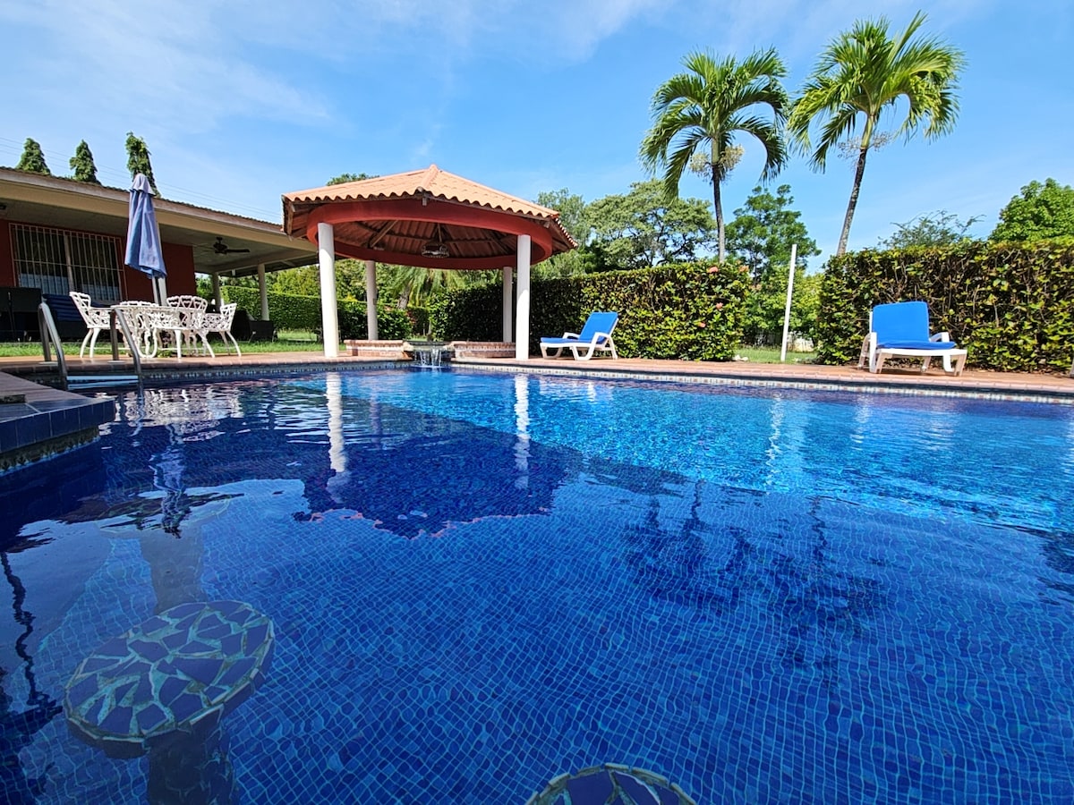 A serene swimming pool is framed by lush greenery and features a gazebo with a tiled roof. Comfortable lounge chairs are situated around the pool, inviting relaxation under clear blue skies.