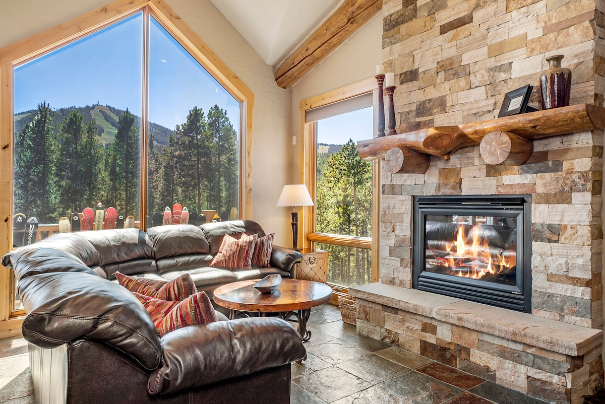 A spacious living area features a large leather sectional couch arranged around a coffee table. A stone fireplace provides warmth, while floor-to-ceiling windows offer views of the mountains and surrounding trees. A lamp stands beside the seating area, adding to the cozy ambiance.
